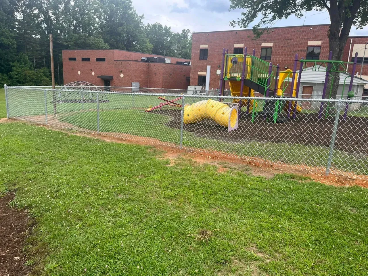 Chain-link fenced playground beside a brick school building, with green grass and yellow play tunnel equipment.