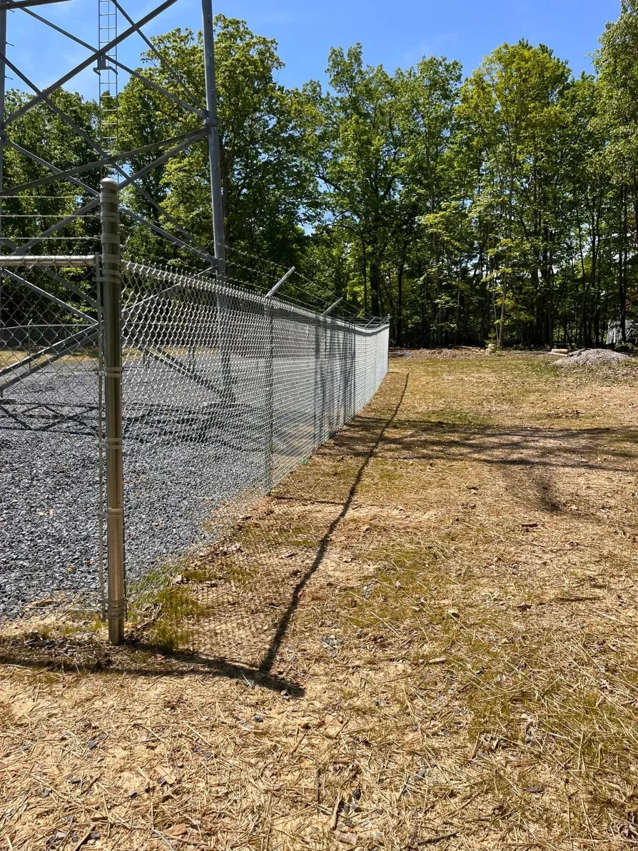 Chain-link fence beside a dry grassy field under trees on a sunny day