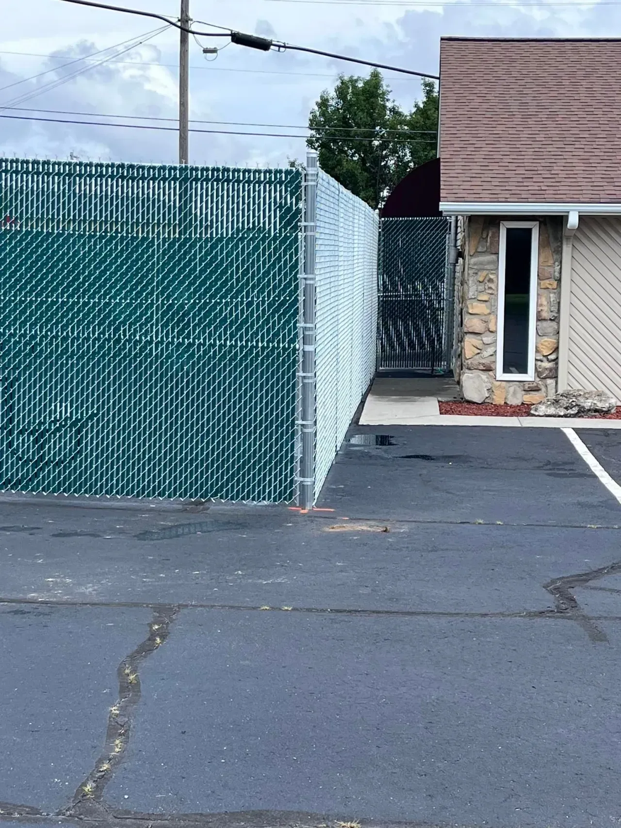 Green chain-link fence beside a paved lot next to a small tan building with a doorway and narrow window