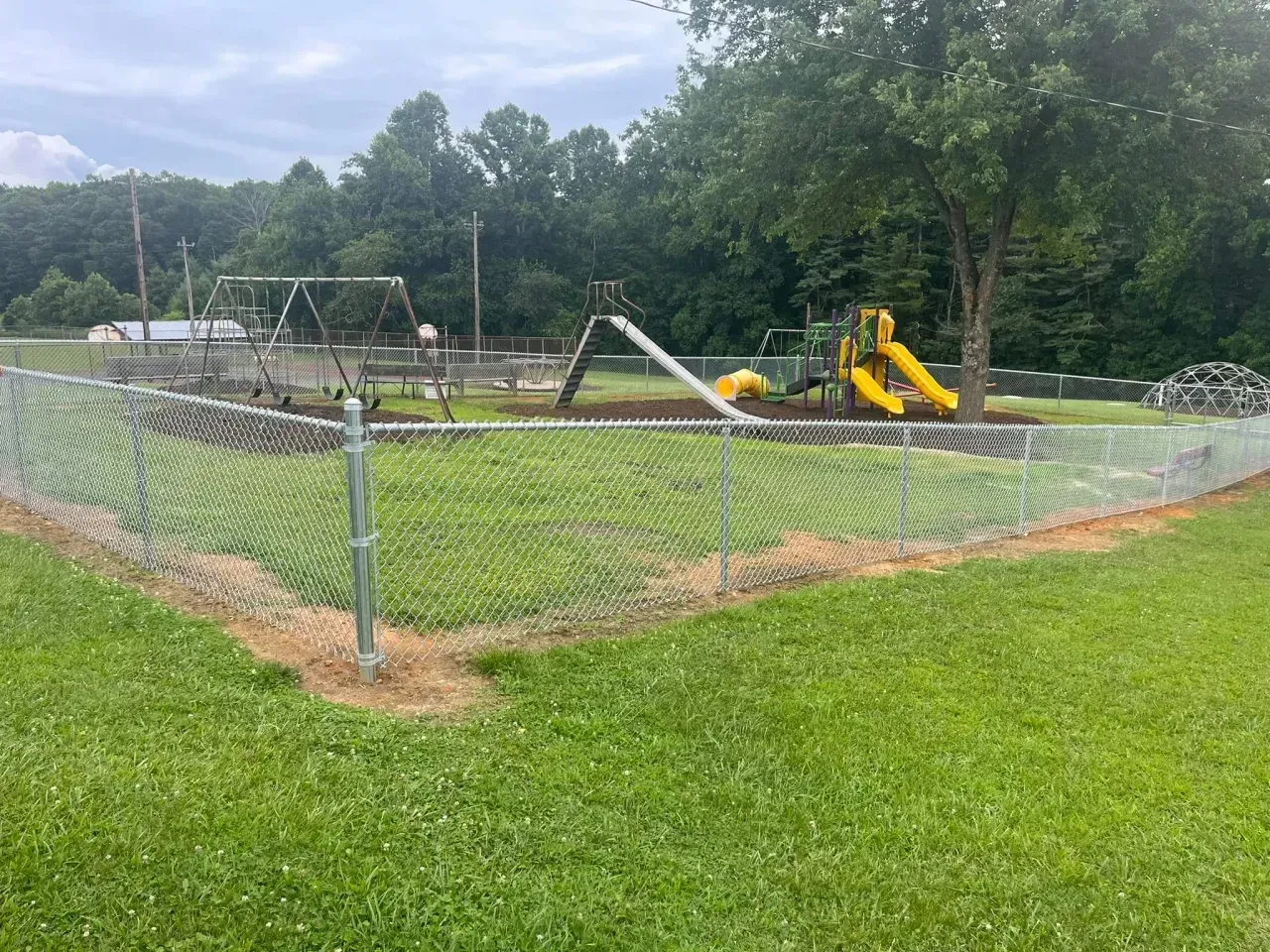 Fenced playground with swings, a slide, and grassy field under cloudy sky