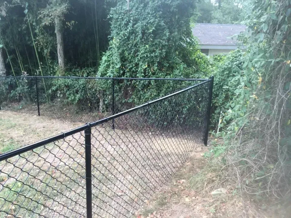 Black chain-link fence beside a grassy, tree-lined yard and house in the background