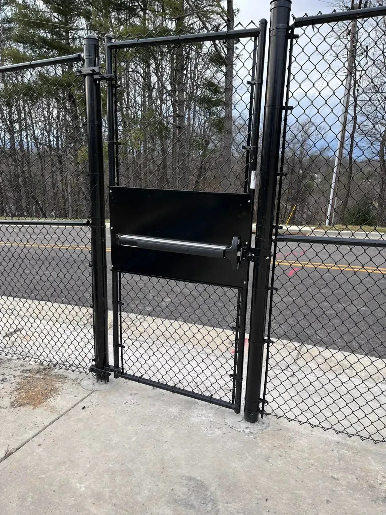 Chain-link fence gate with a black privacy panel beside a road and bare trees