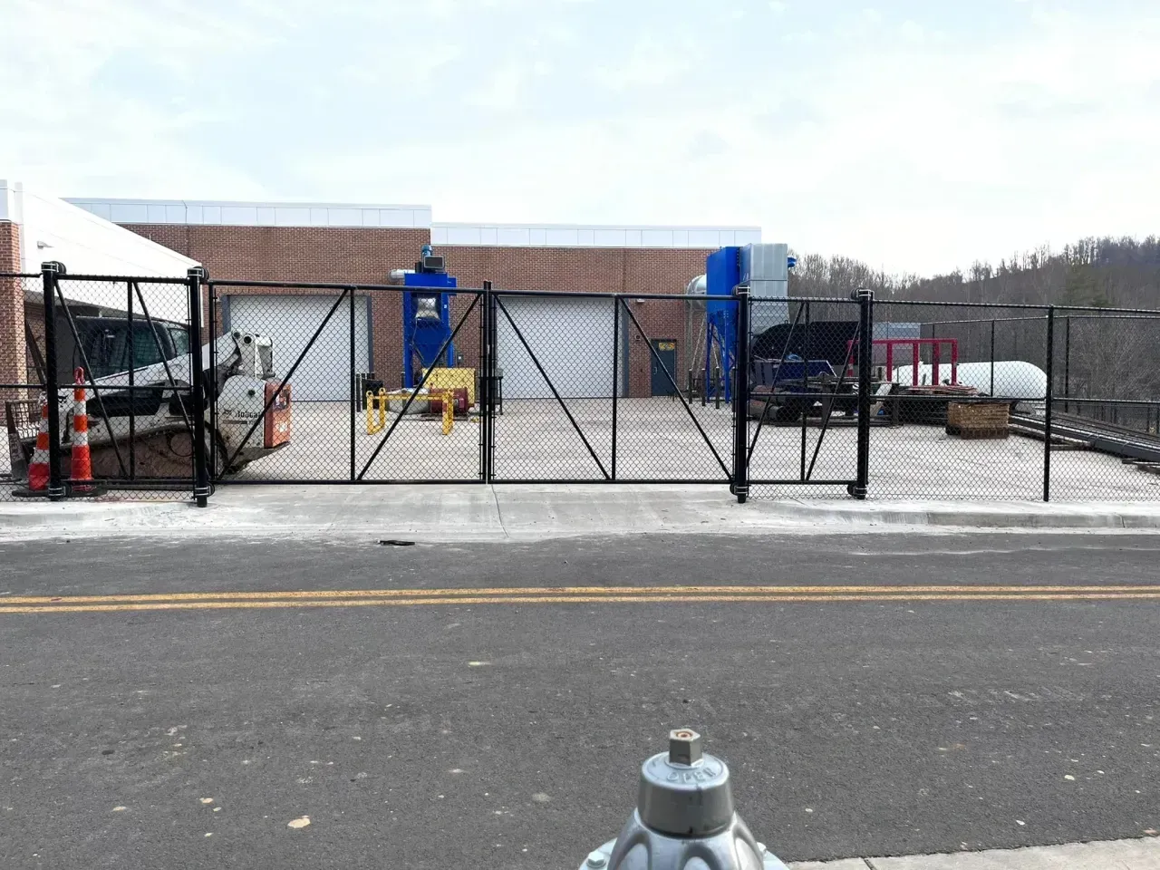 Snowy industrial yard with a black chain-link gate, blue machinery, and a fire hydrant in the foreground
