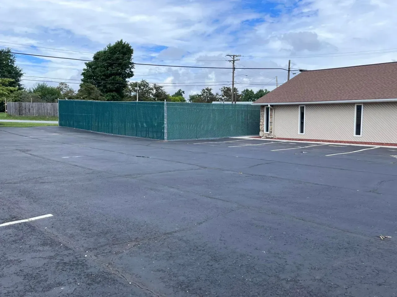 Empty parking lot beside a beige building and green fence under cloudy sky