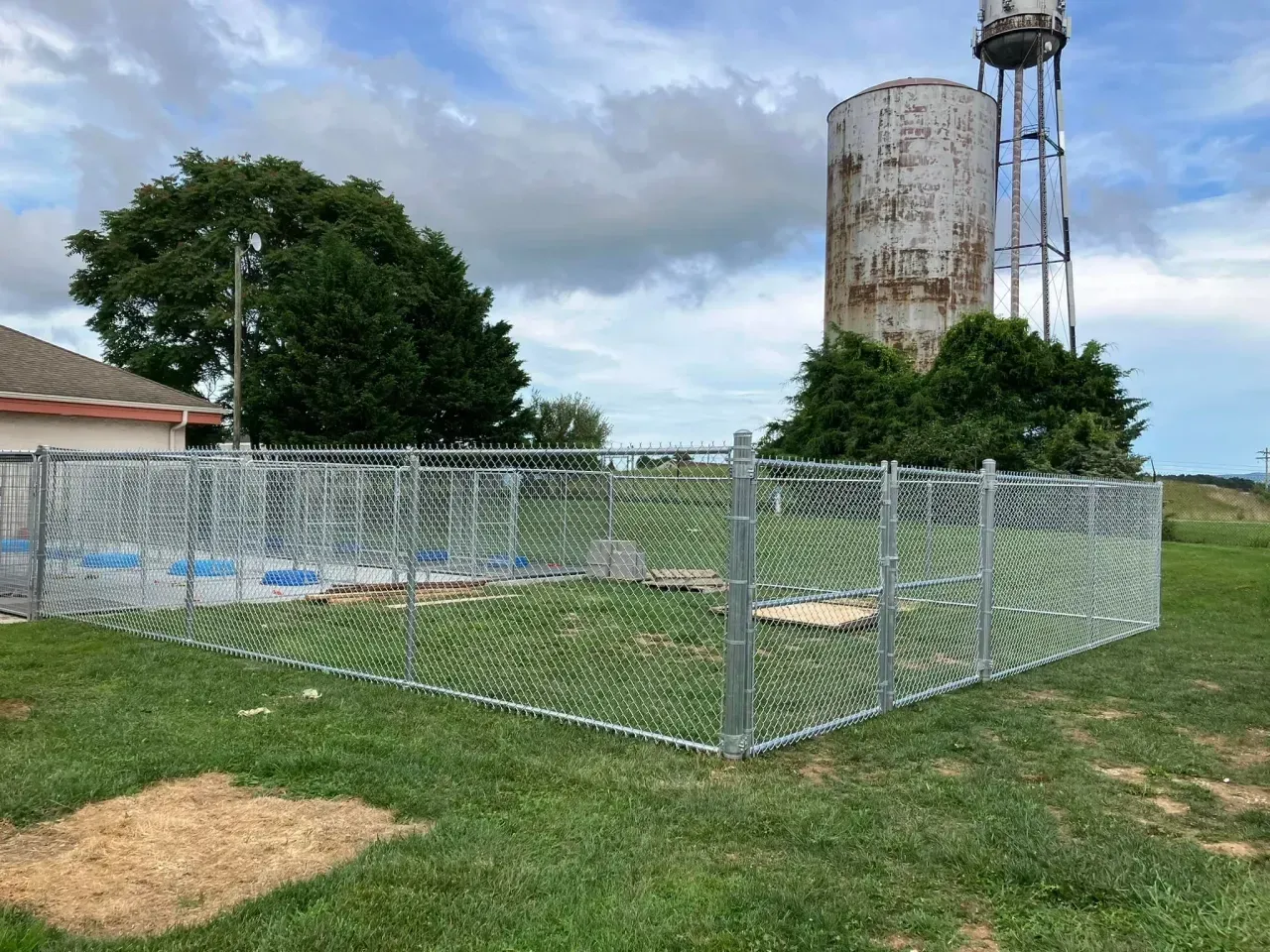 Chain-link fenced area beside a rusty water tower in a grassy field under cloudy sky