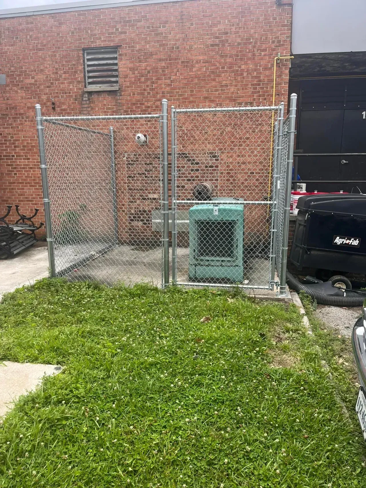 Chain-link fenced utility area beside brick building with green trash bin and black dumpster on pavement.