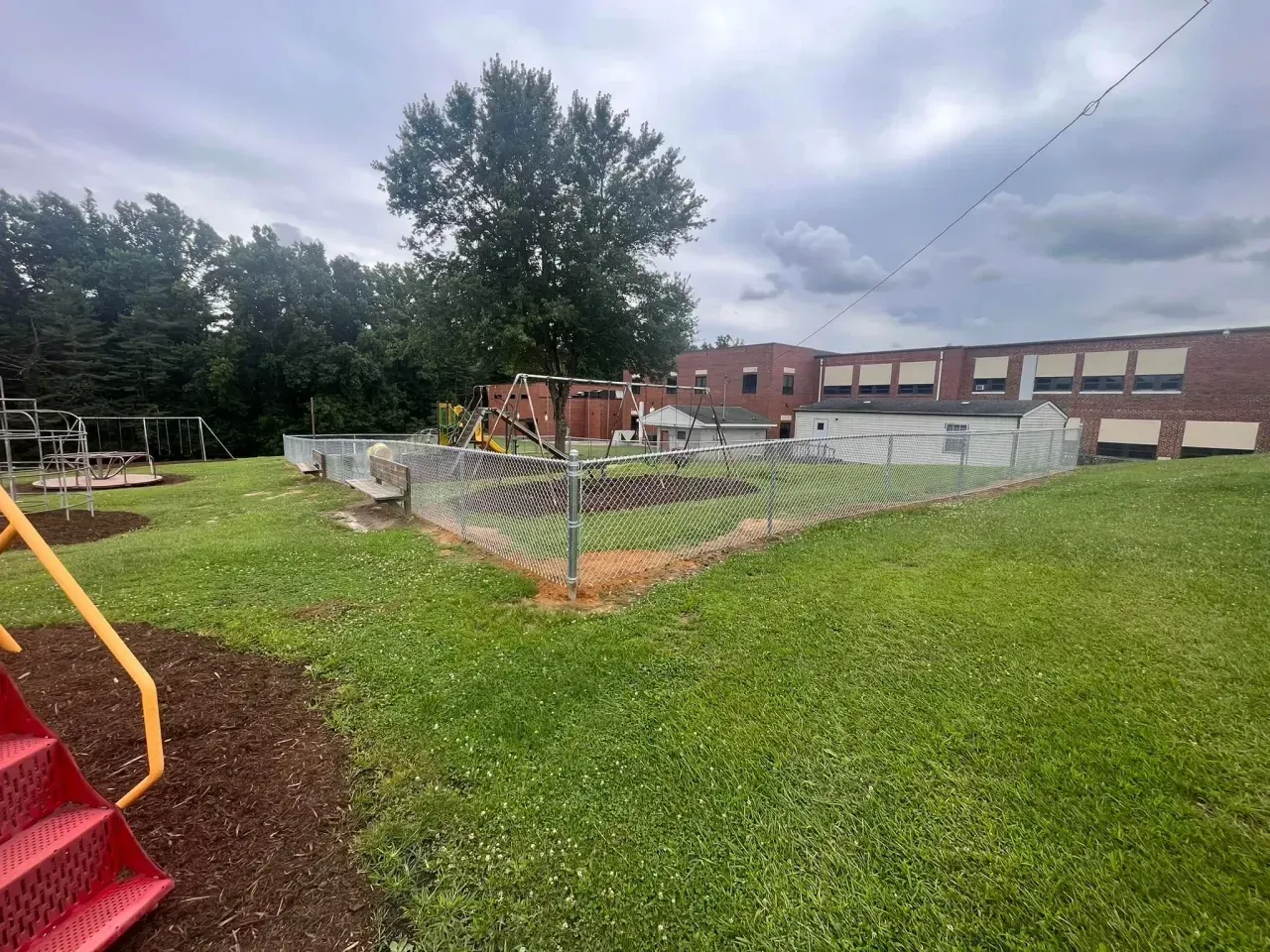 Fenced school garden beside a brick building, with green grass and cloudy sky