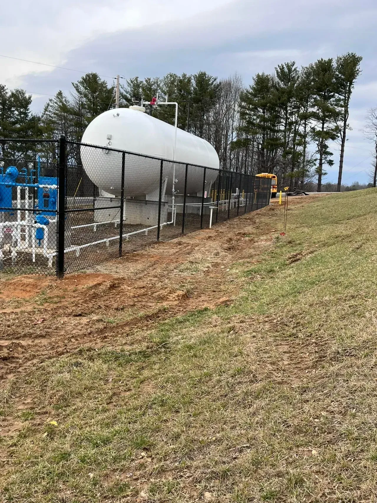 White cylindrical tank behind a black fence on a grassy slope, with trees and utility equipment nearby