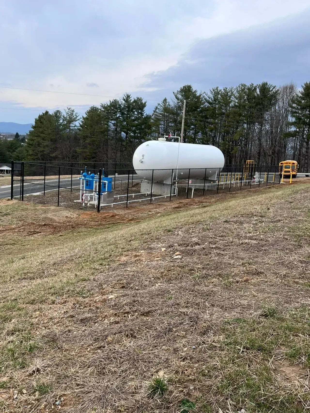 White propane tank in a fenced yard with blue equipment and trees under a cloudy sky