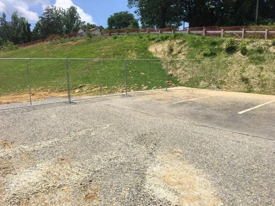Empty gravel lot beside a chain-link fence and grassy hillside under a clear sky.
