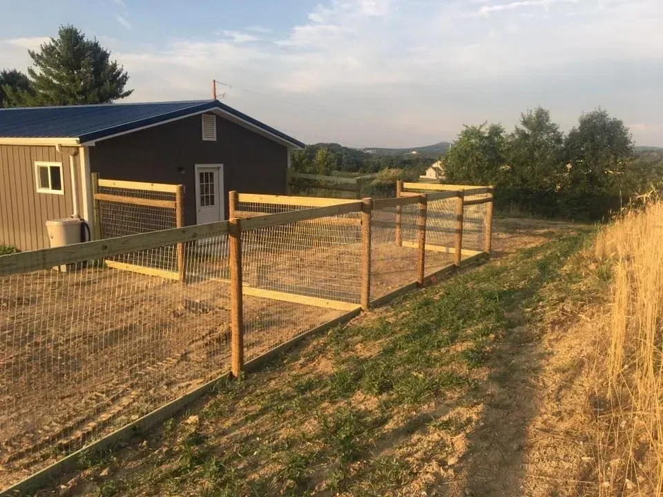 Fenced yard beside a small brown house at sunset, with dry grass and distant hills.