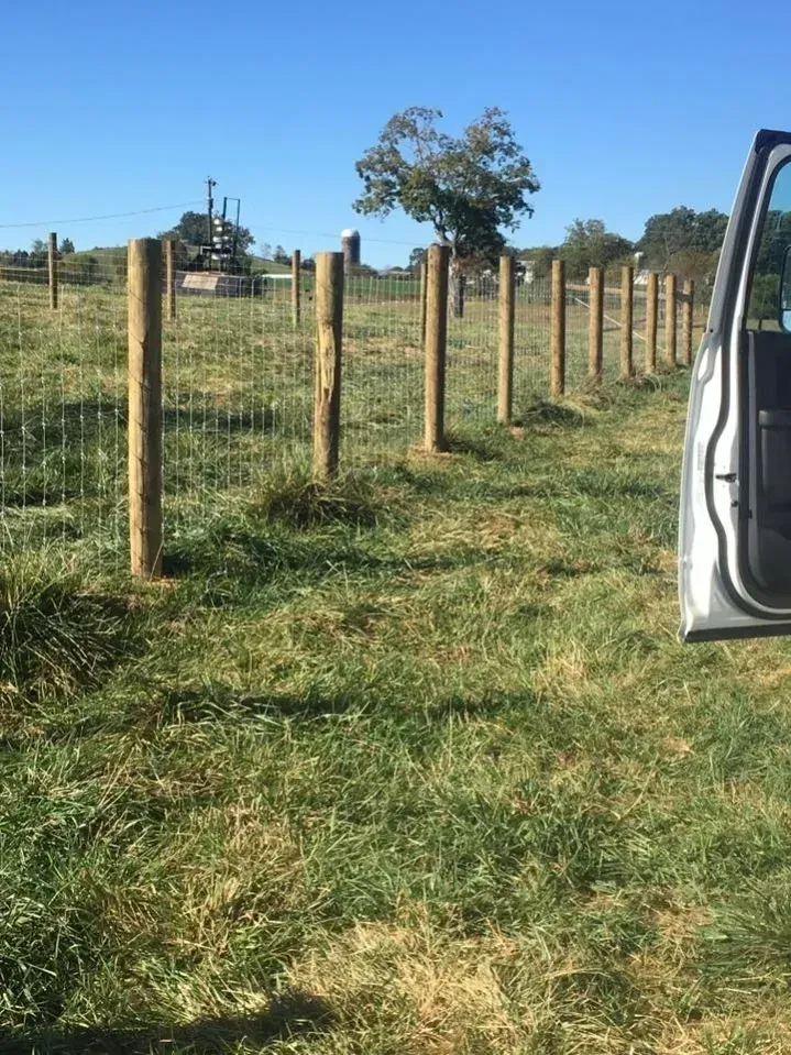 Wooden fence posts lining a grassy field under a clear blue sky, with an open vehicle door at right.