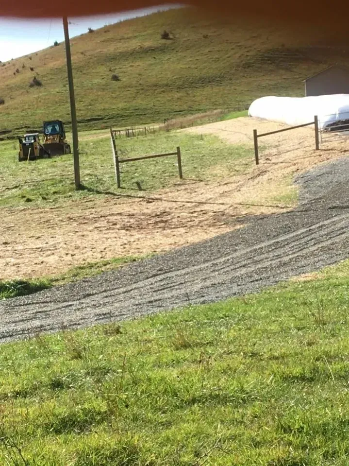 Rural field with a small tractor, fenced gate, gravel path, and white tank near a grassy hill