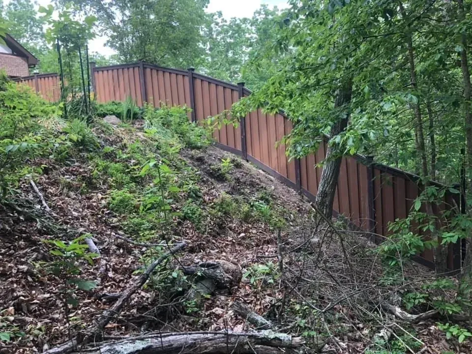 Wooded hillside with a brown wooden fence along the top and dense green trees.