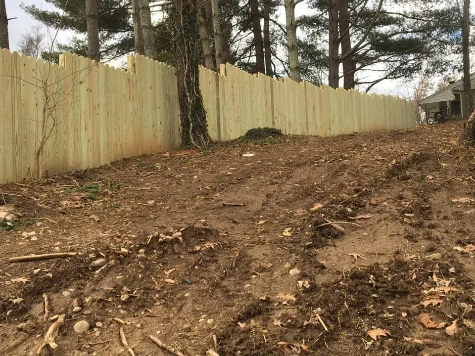 Dirt slope beside a pale wooden fence with trees and a small shed in the background.