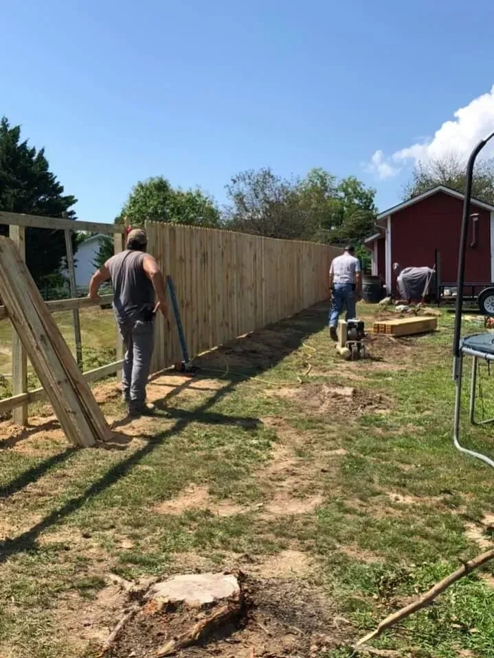 Two people building a wooden fence in a sunny backyard beside a red shed