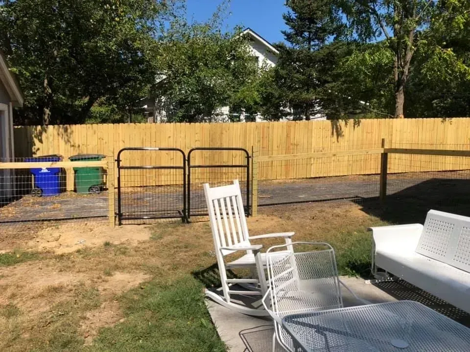 Backyard with a wooden fence, metal gate, and white patio chairs in the foreground.
