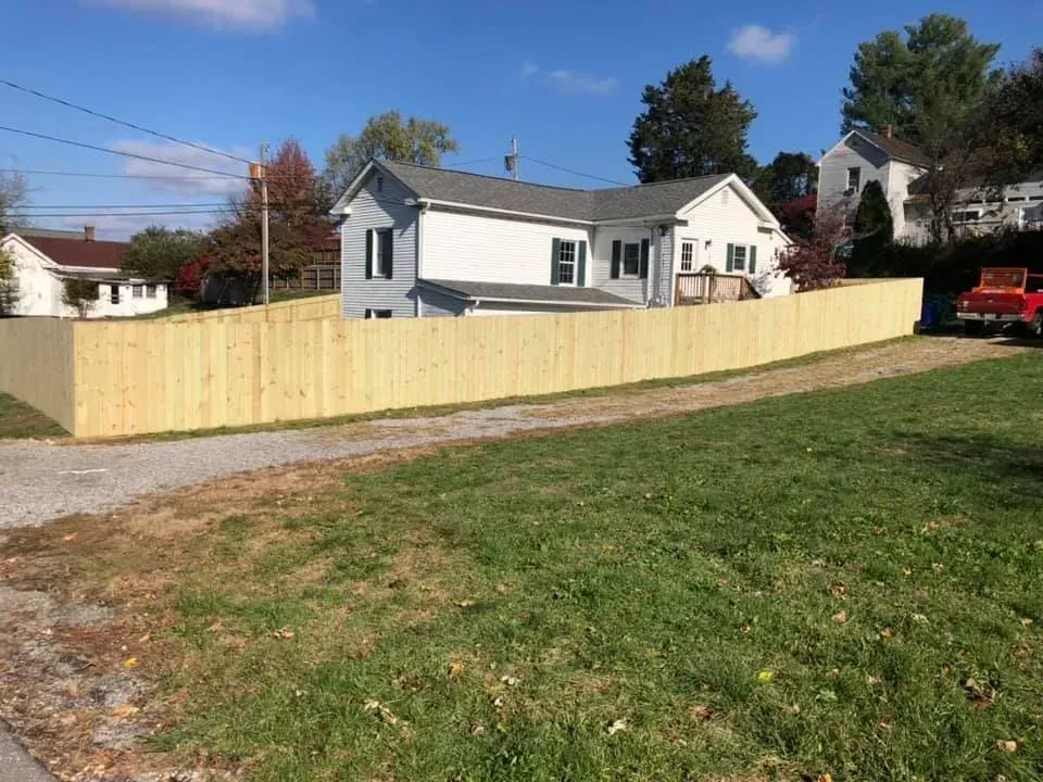 White house behind a long tan wooden fence beside a grassy yard and gravel drive under a blue sky