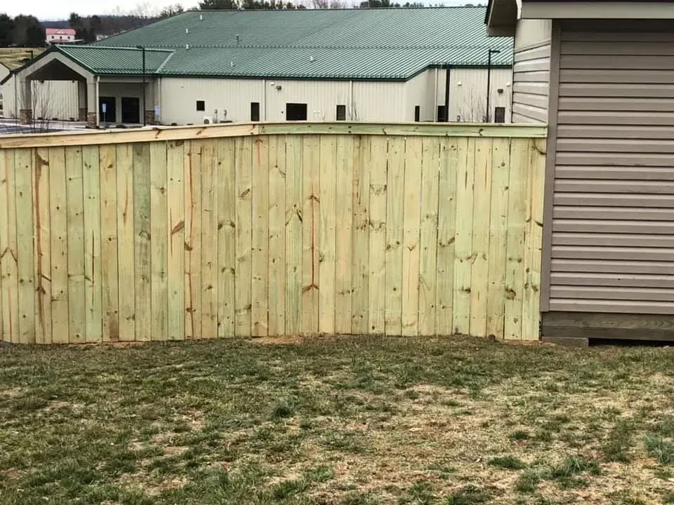 Light wooden fence in a yard beside a beige building with a green roof and patchy grass foreground