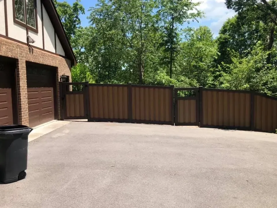 Driveway beside a brick house with a brown garage and matching privacy fence, backed by trees.