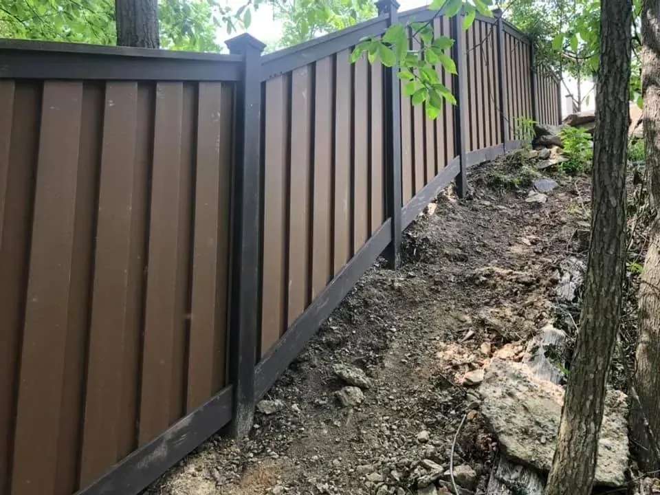 Brown wooden fence along a sloped dirt embankment beside trees and rocks