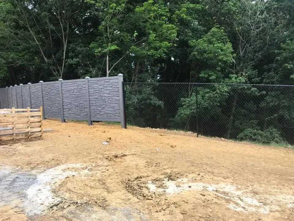 Dirt lot beside a gray fence and chain-link fence with trees in the background