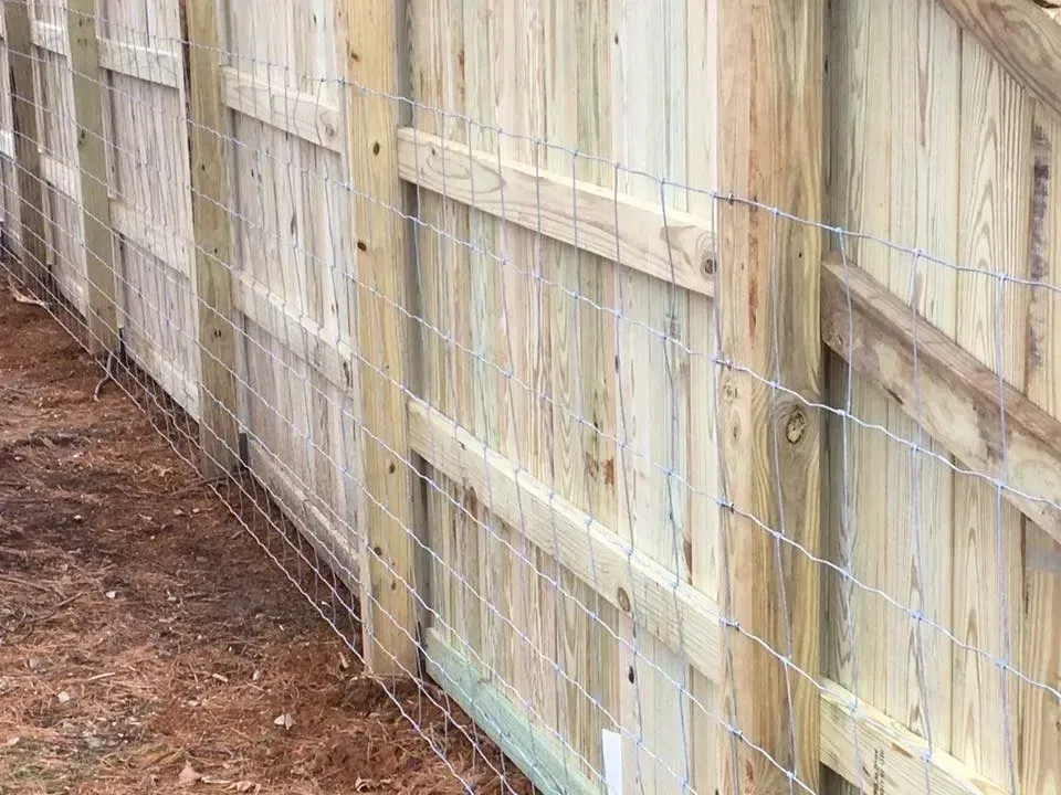 Wooden privacy fence along a dirt path, with wire mesh reinforcement and light-colored panels.