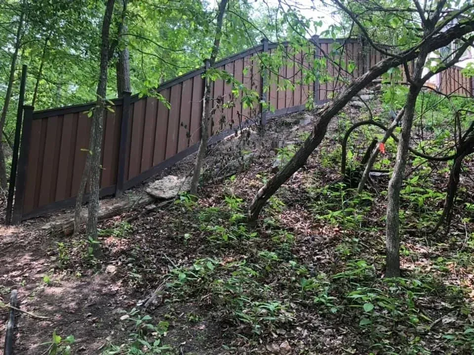 Brown wooden fence on a steep wooded slope with a dirt path and trees around it