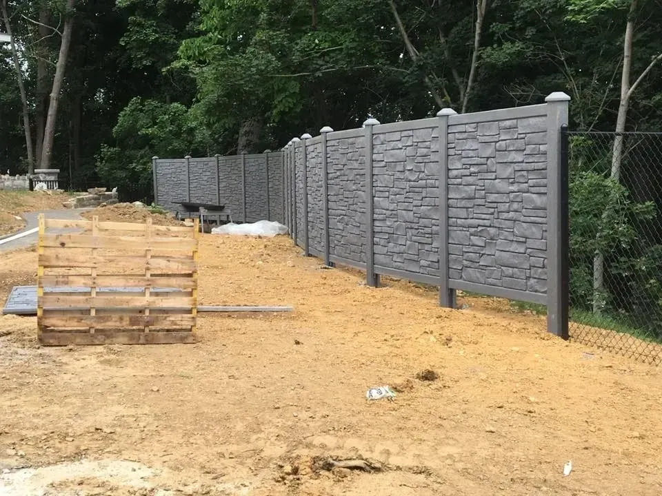 Gray stone fence under construction beside a wooded area, with stacked lumber and dirt ground.