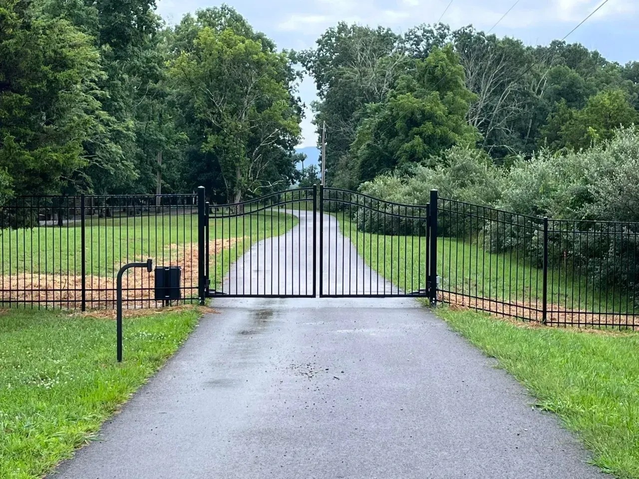 Closed black gate across a paved driveway, flanked by green grass and trees in a wooded area