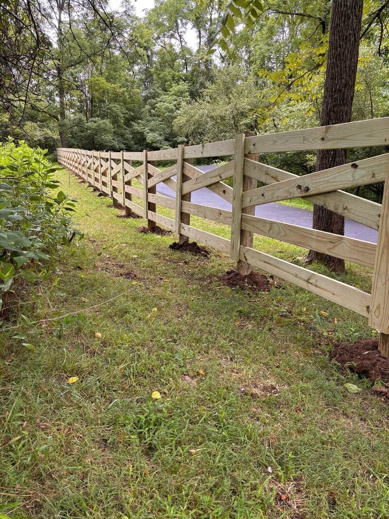White wooden fence along a wooded roadside with grass and trees.