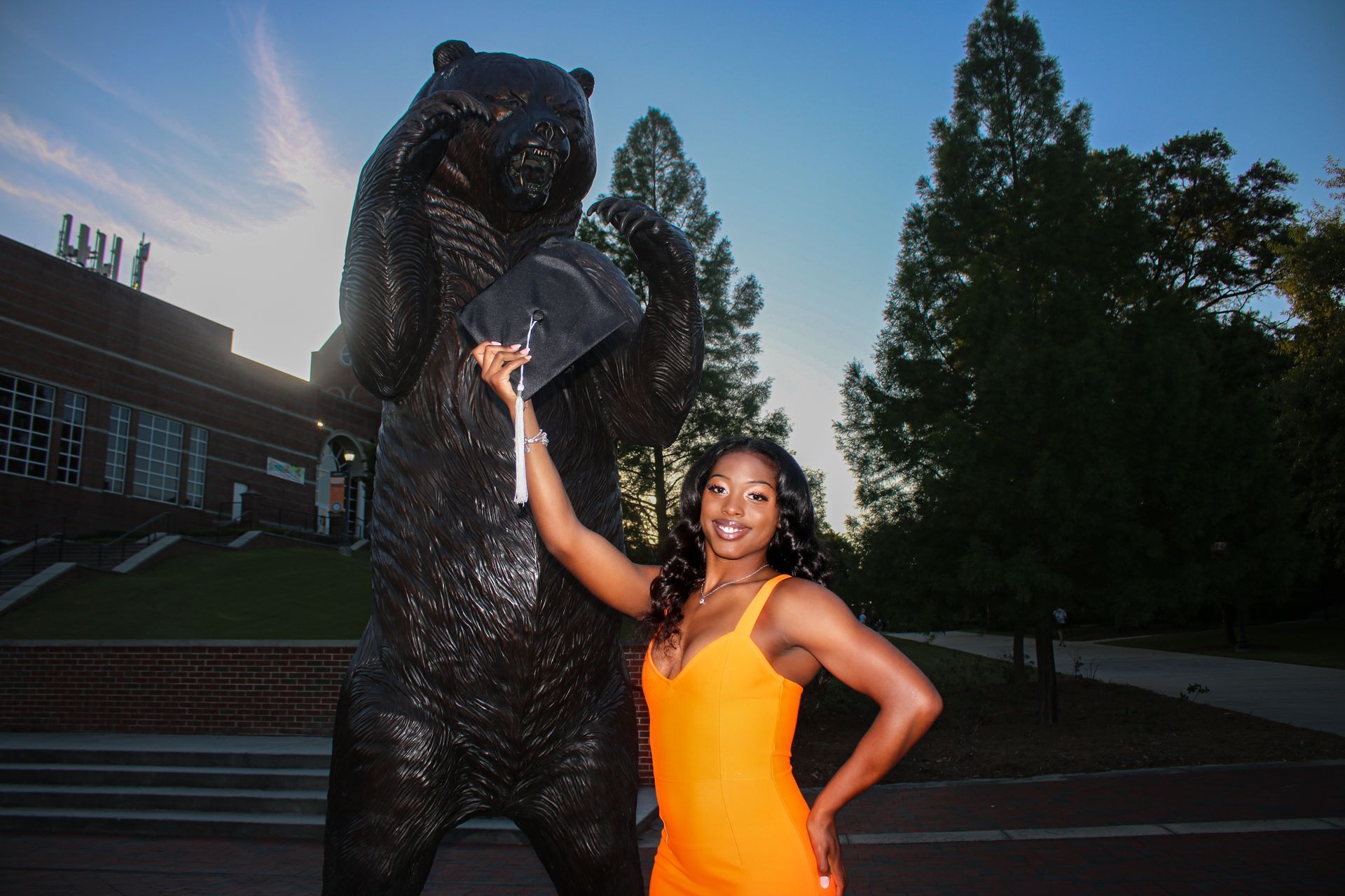 Woman in orange dress posing beside a large black bear statue outdoors at dusk