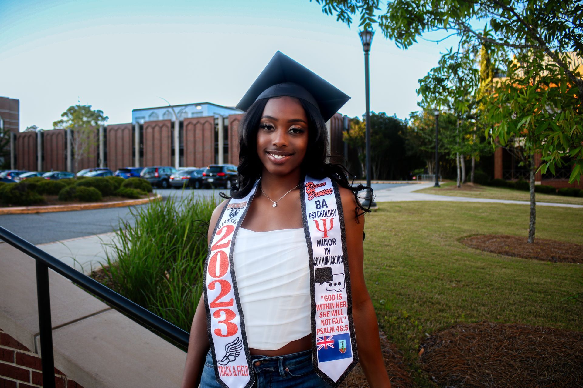 Graduate in cap and stole smiles outdoors on a campus walkway, with a brick building in the background.