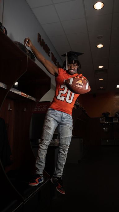 Person in an orange sports jersey holding a football in a locker room, arms raised.