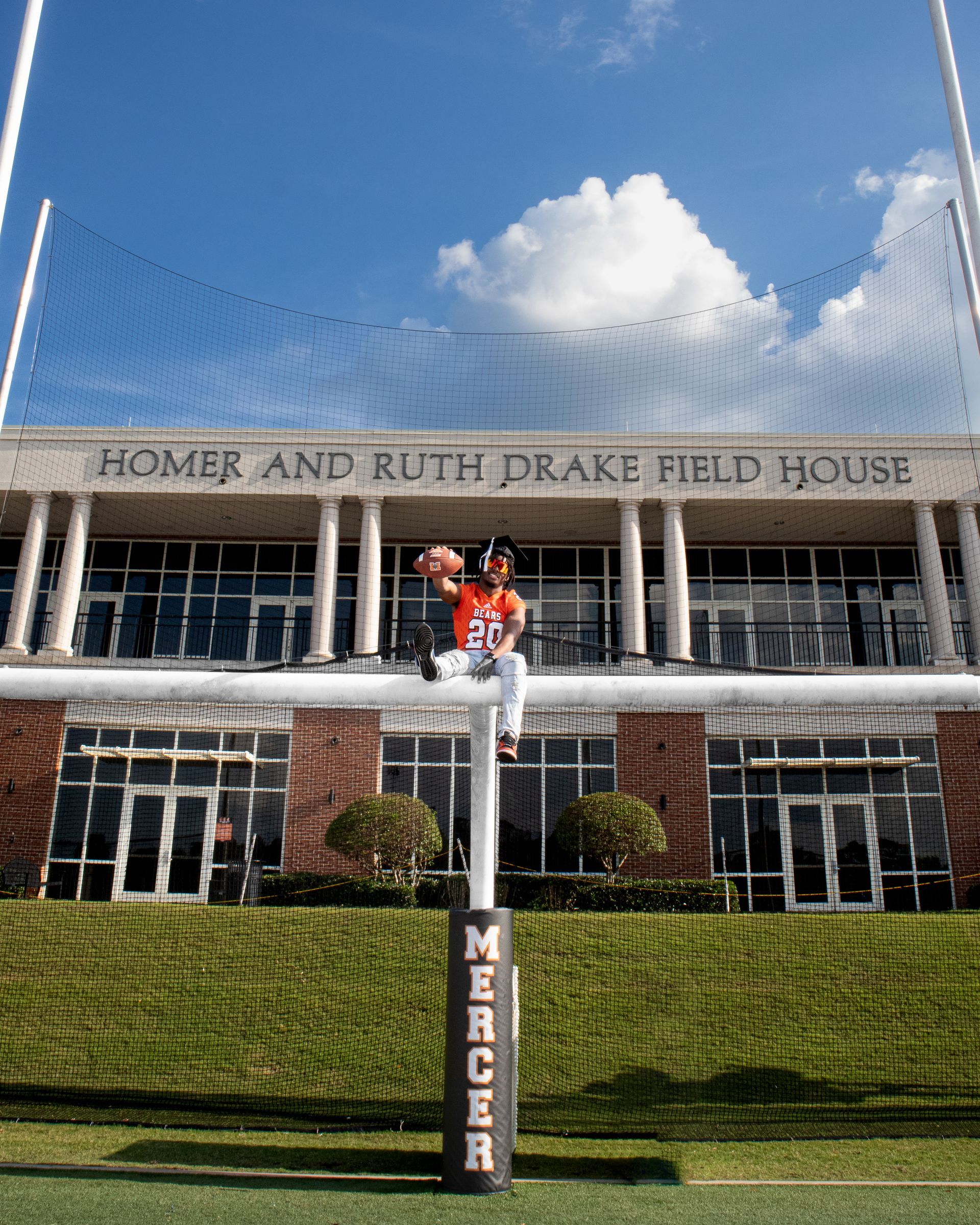 Home plate at Drake Field House with a large building facade, goalpost, and bright blue sky with clouds.