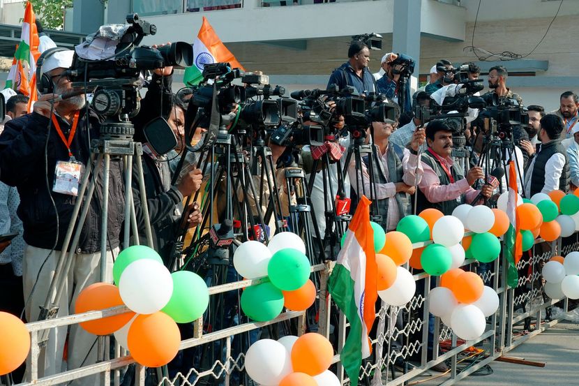 Cameras and reporters behind tricolor balloon decorations at a crowded outdoor event