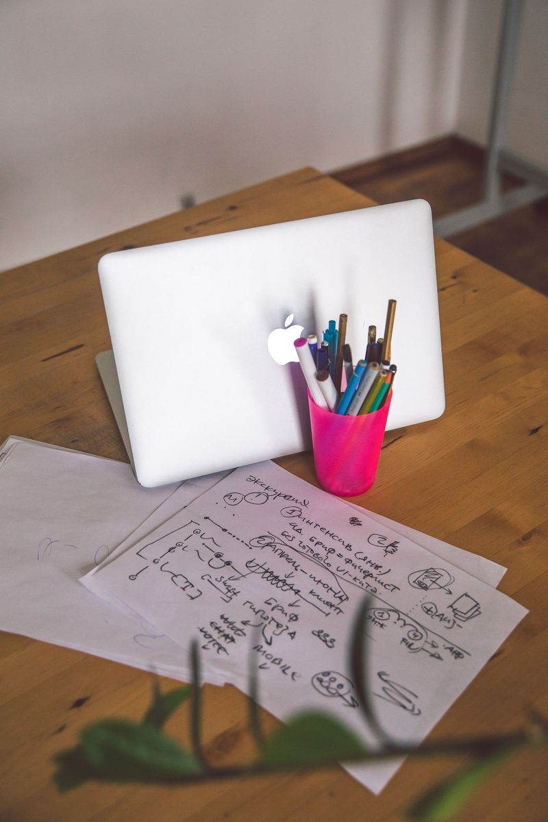 White laptop, pink pen cup, and papers on a wooden desk with a green plant.