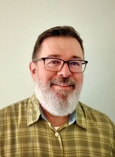 Man with glasses and grey beard smiling in front of a light green wall, wearing a plaid shirt.