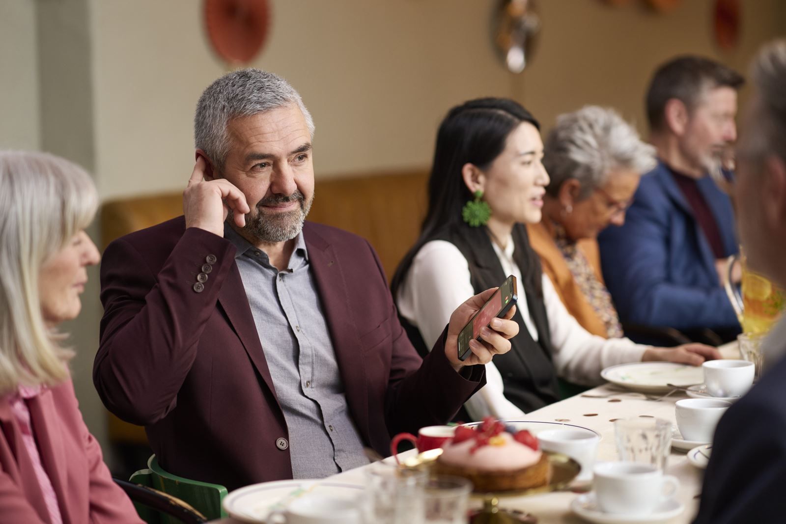 Man in burgundy jacket holds phone, appears to listen while sitting at a table with others.