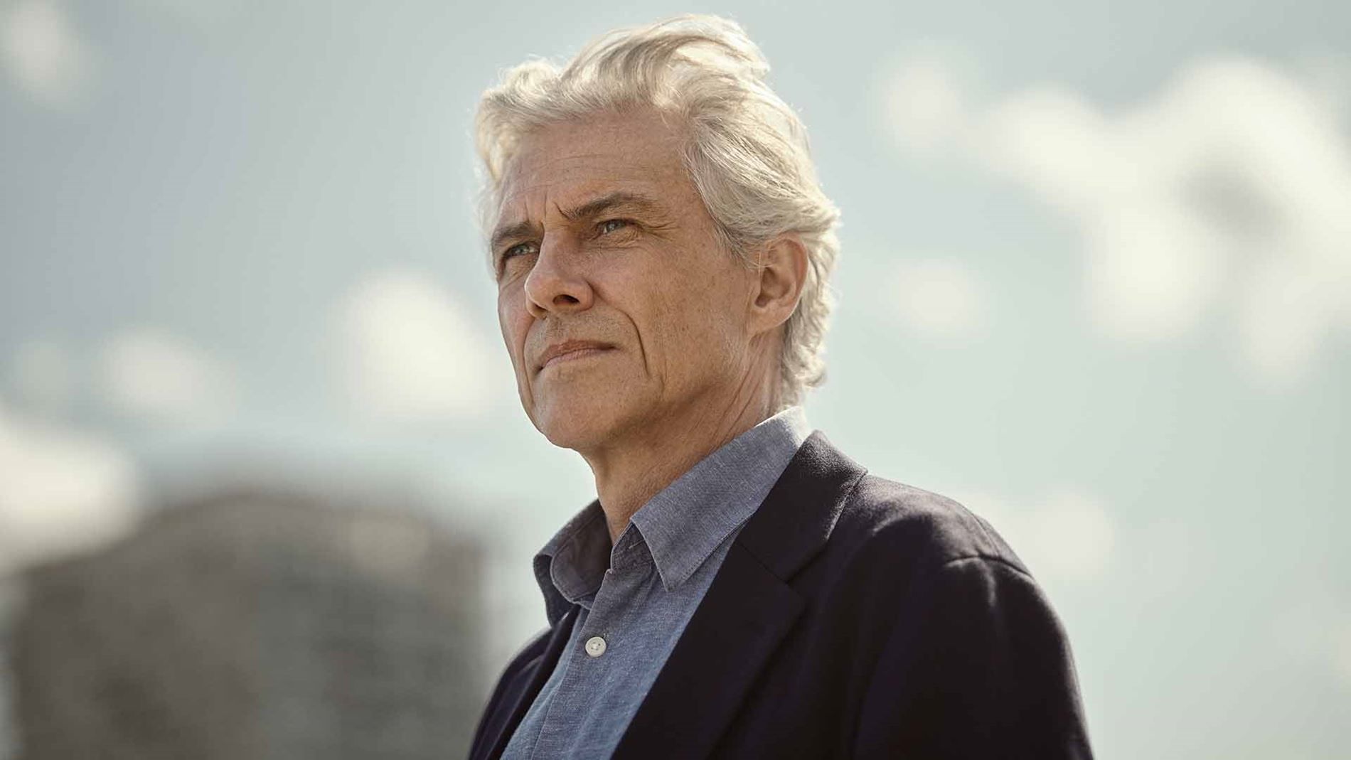 Man with gray hair looks up, wearing a blue shirt and navy blazer; city skyline in background.