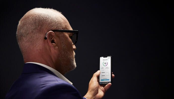 Man in suit looking at phone screen, wearing glasses and earbud, dark background.