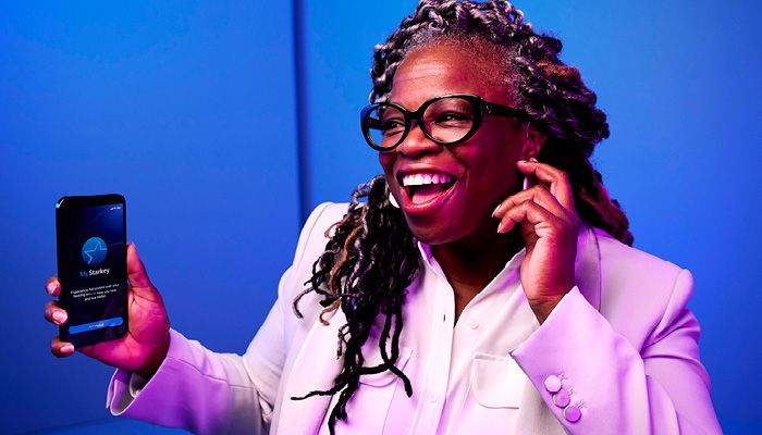 Woman holding phone, smiling, wearing glasses and blazer in blue-lit setting. Phone screen visible.