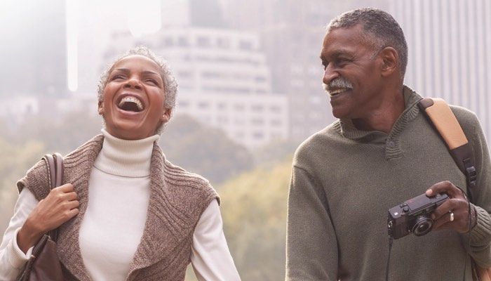 Smiling couple walking outdoors, woman laughing, man holding camera, cityscape background.