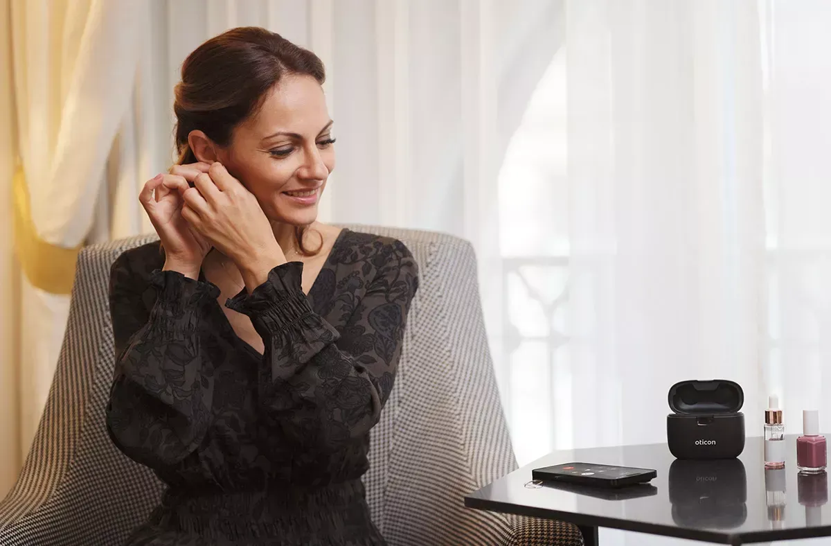 Woman putting on earrings, sitting by a window. Black dress, neutral-toned chair. Small table with earbuds.