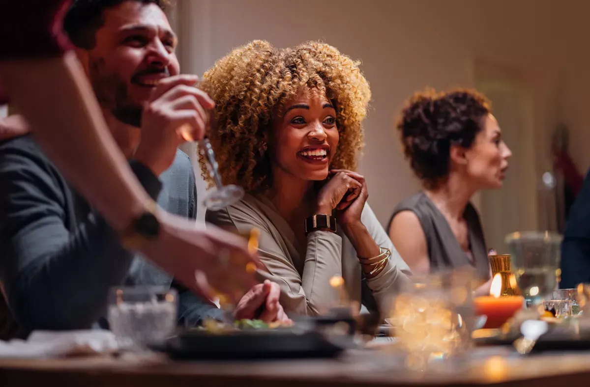 Friends at a dinner table, smiling. Warm lighting, food and drinks present.