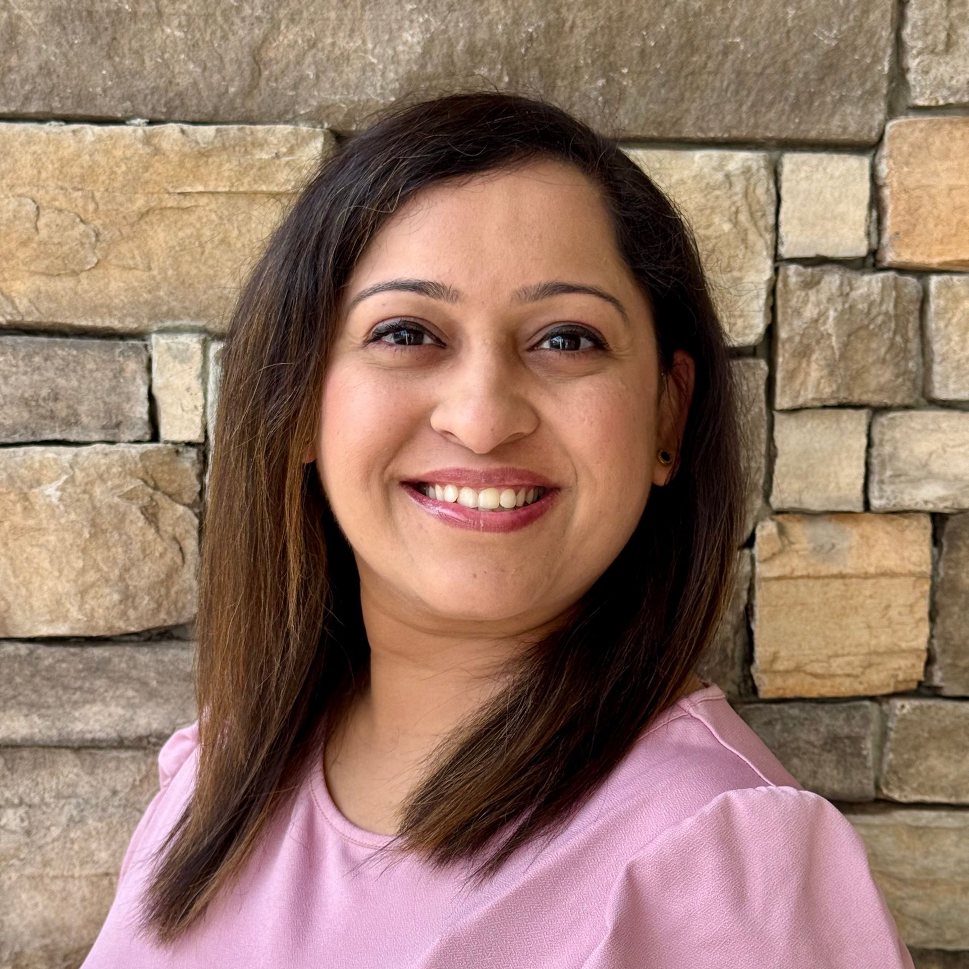 A smiling person with dark hair wearing a light pink top, positioned against a background of stacked tan stone.