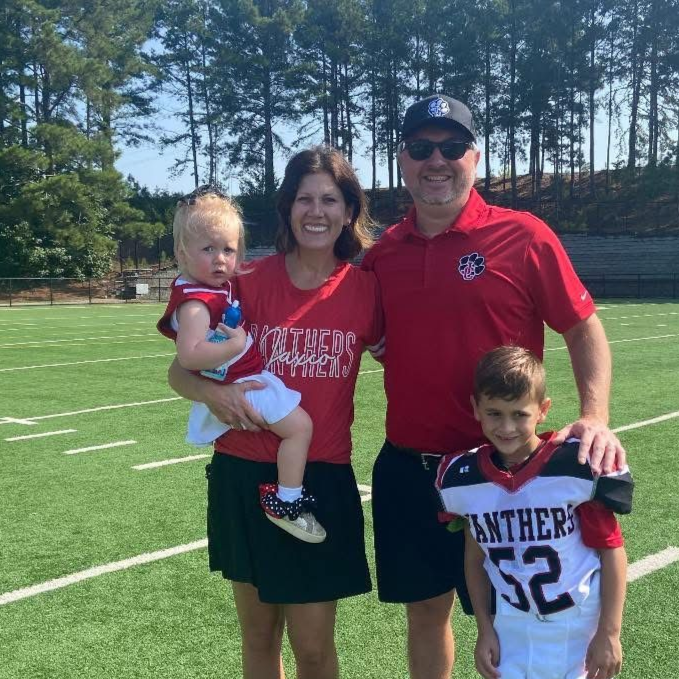 A family stands on a grassy sports field. A woman holds a young child; a man stands beside a boy wearing a team jersey.