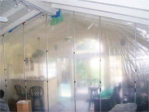 Transparent plastic sheeting encloses a room with chairs, boxes, and hanging tools near the ceiling.