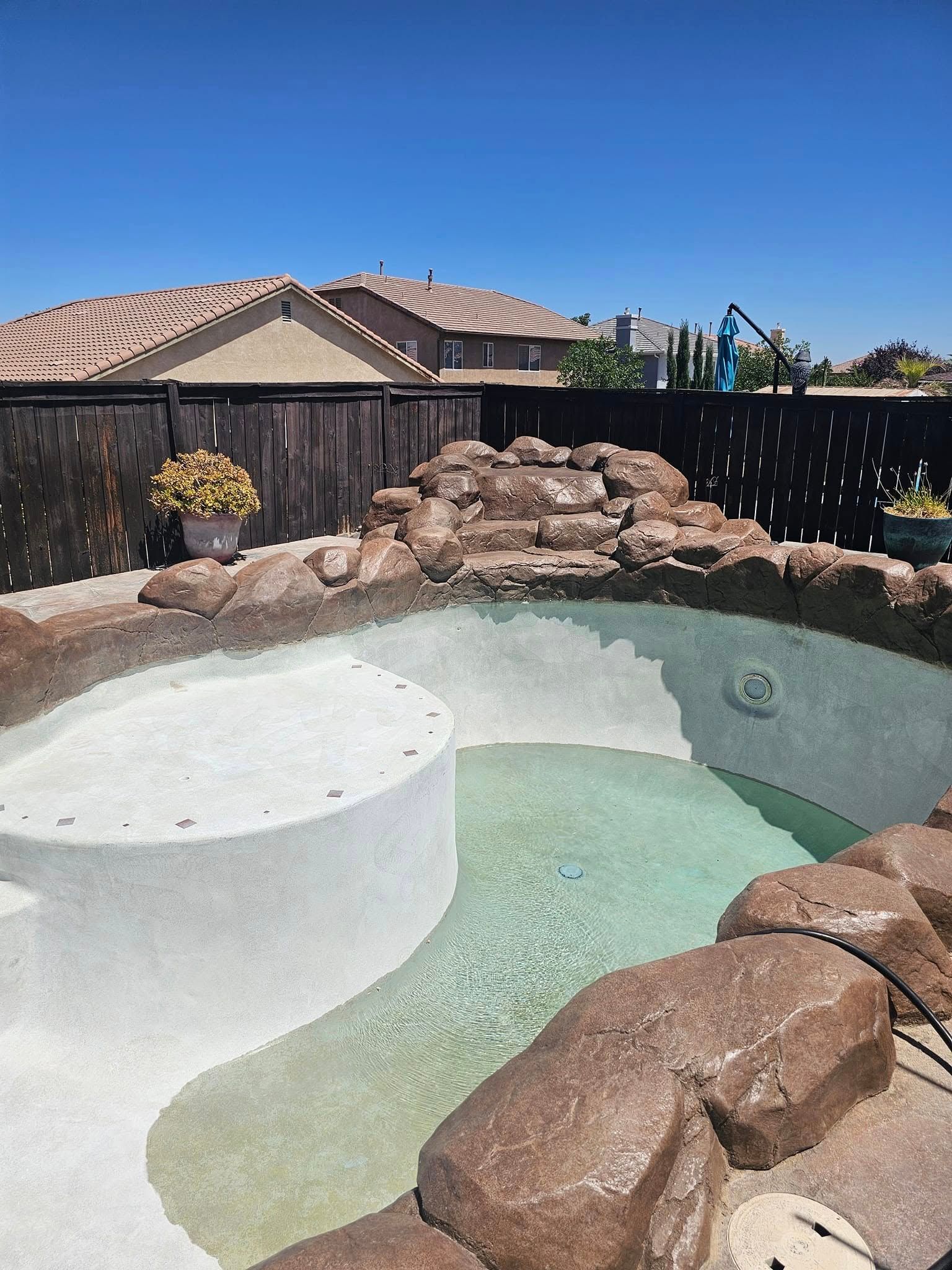 Backyard swimming pool with a white spa, rock waterfall, and desert-style fencing under a clear blue sky