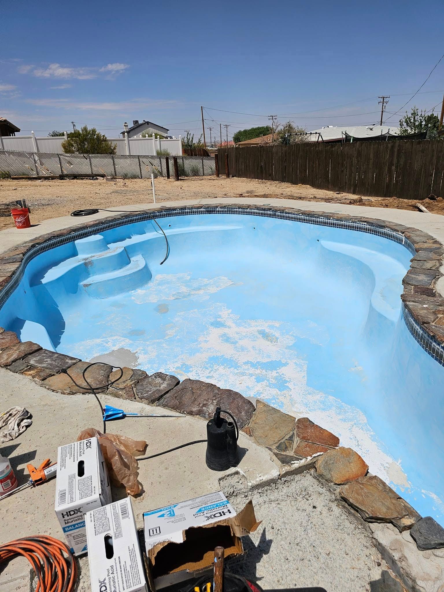 Empty backyard pool with blue interior, rock border, and patio tools in the foreground under a sunny sky